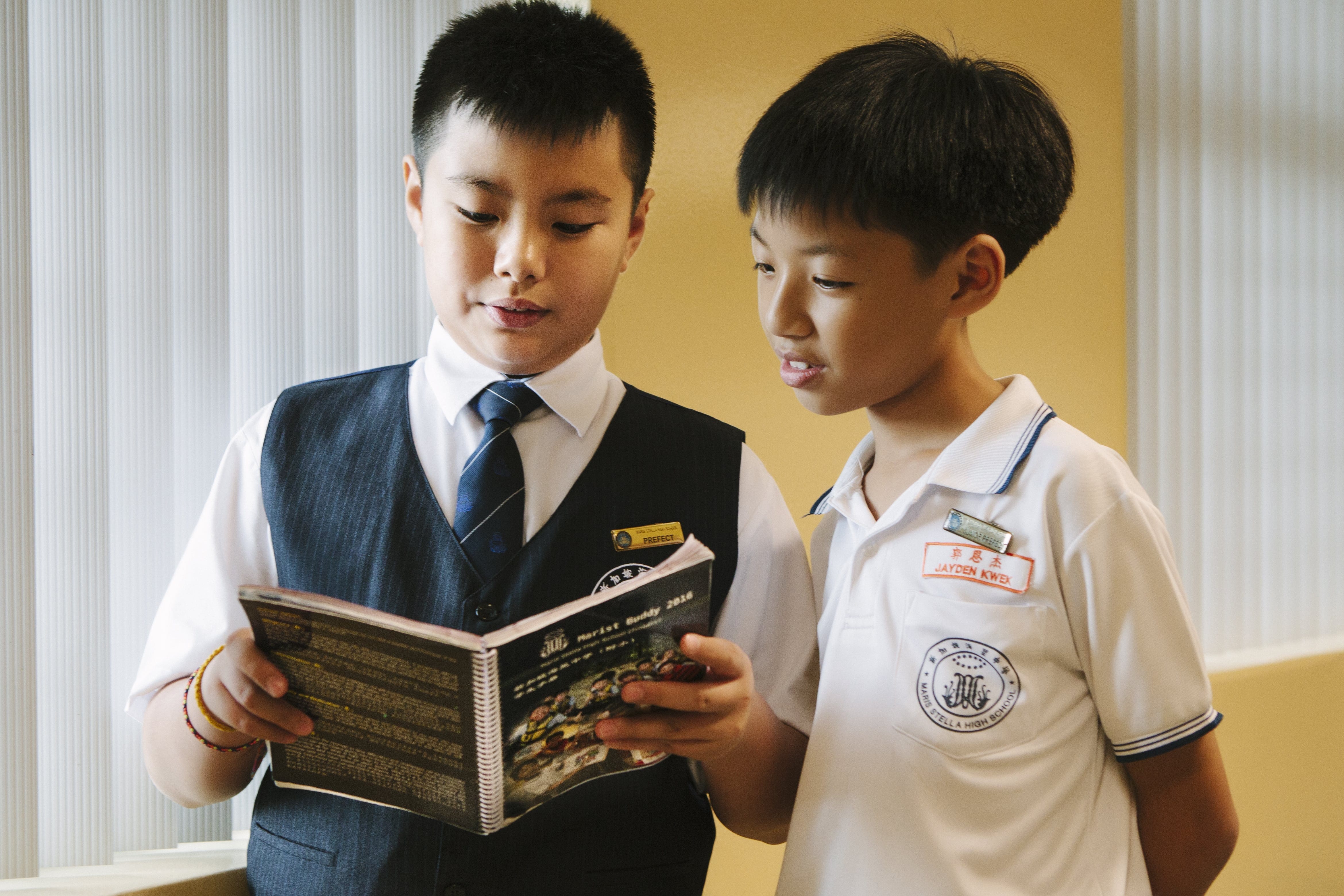 Two boys in school uniforms reading a spiral-bound booklet together indoors.